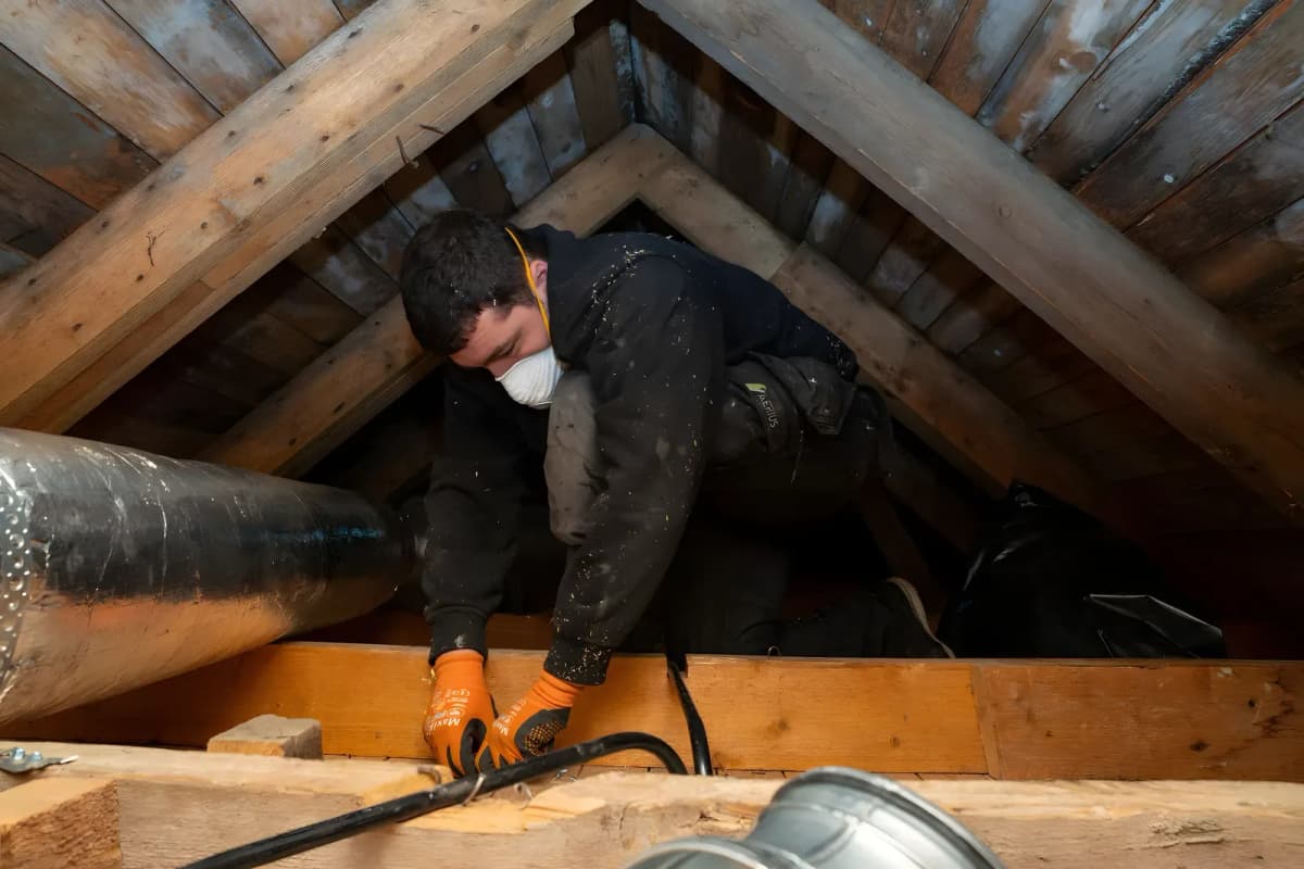 Tekniker med andningsskydd och orange handskar arbetar med en kabel på en vind bredvid isolerad ventilationskanal