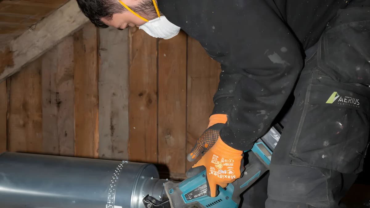Tekniker med munskydd och orange handskar arbetar med en galvaniserad ventilationskanal på vinden i Stockholm